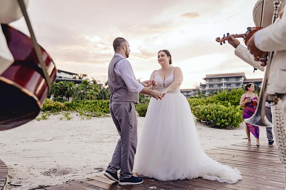 Mariachi band plays music while bride and groom dance beachfront.