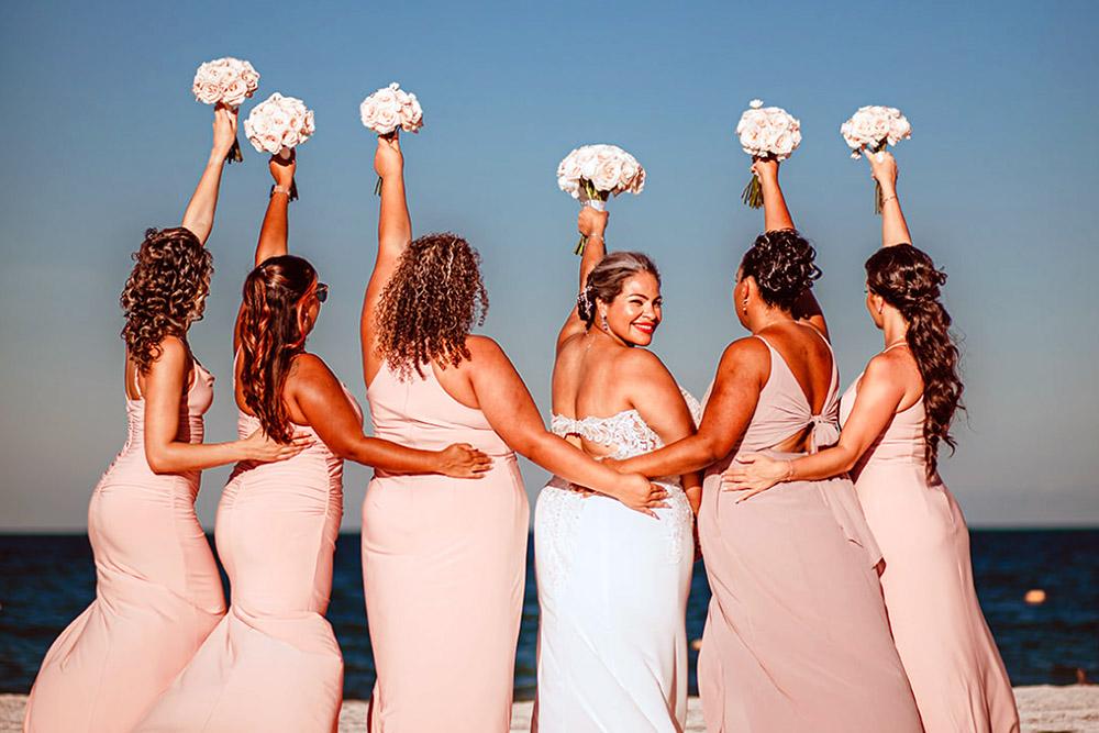 Bride and bridesmaids on a sunny beach holding their bouquet of flowers in the air.