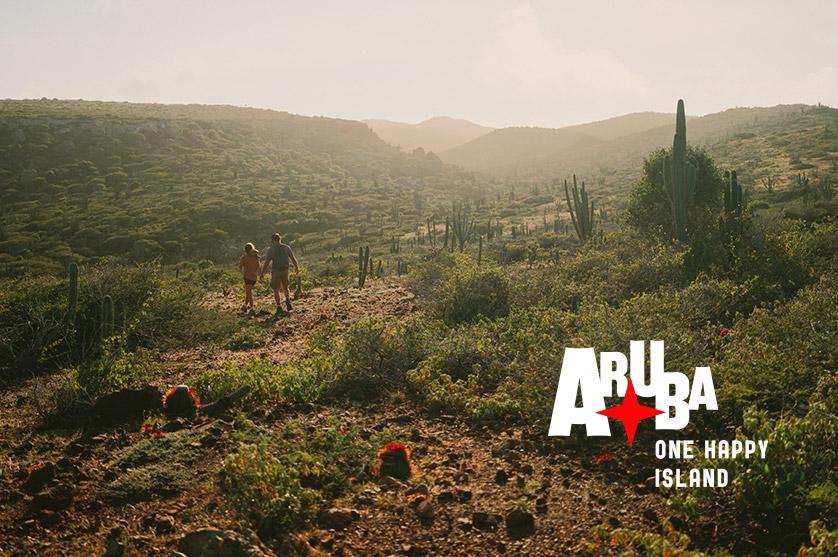 Two hikers explore the rugged terrain of Aruba, walking along a dusty path surrounded by various cacti and scrub brush under a soft, hazy sunlight. The "Aruba: One Happy Island" logo is visible in the bottom right corner.
