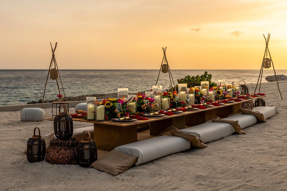 Beach dining setup with a long table, cushions for seating, lanterns, and floral arrangements, set against a sunset ocean backdrop.