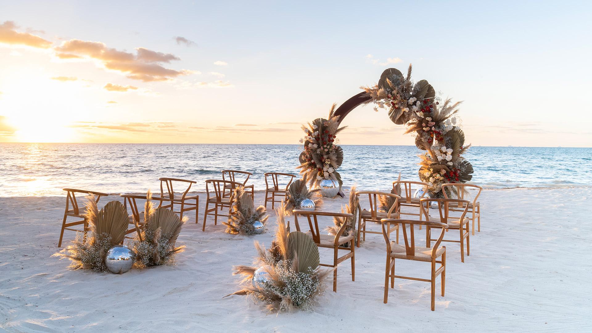 Sunset view of a beach wedding ceremony with wooden chairs, a floral arch, and unique mirrored disco ball decorations.