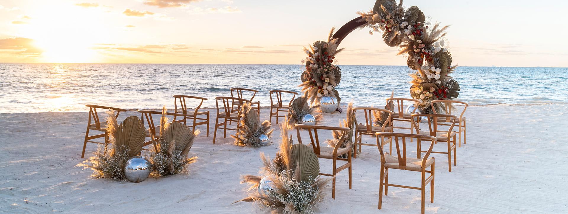 Sunset view of a beach wedding ceremony with wooden chairs, a floral arch, and unique mirrored disco ball decorations.